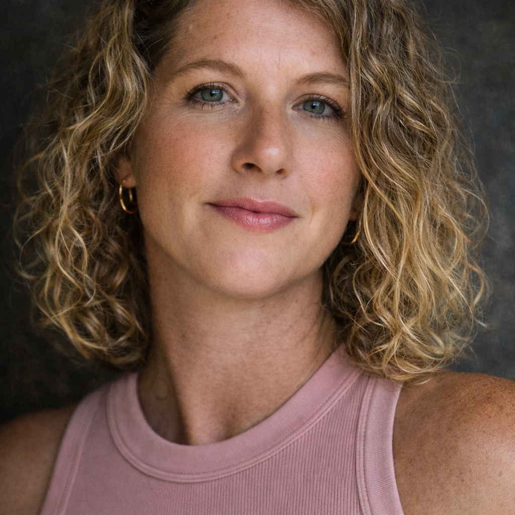 Shailah with curly blonde hair, blue eyes, and light makeup, wearing gold hoop earrings and a pink sleeveless top.