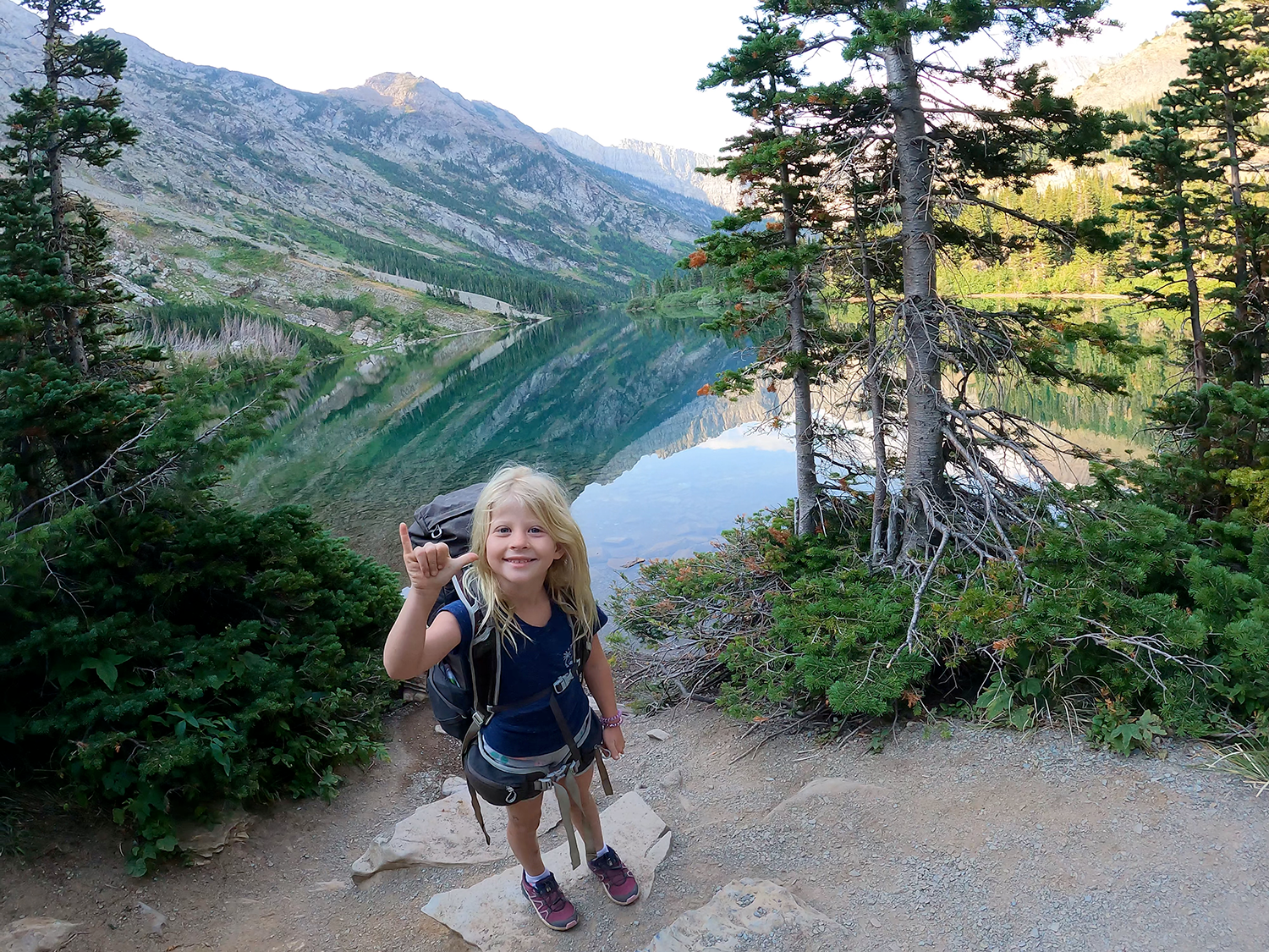 A young girl with blonde hair, wearing a navy shirt, shorts, and sneakers, is smiling and holding up her hand in a shaka sign while hiking near a lake surrounded by mountains and trees.