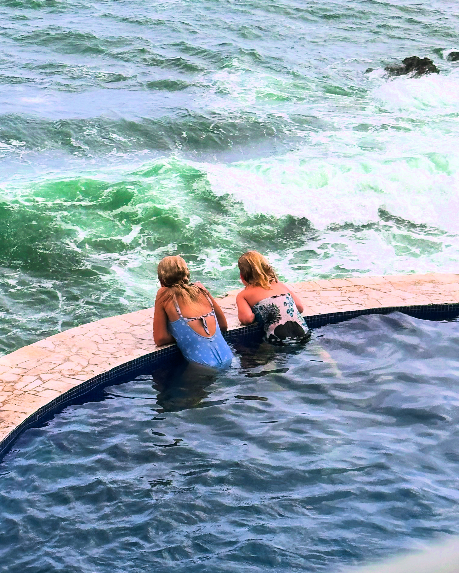 Two girls with blonde hair, wearing swimsuits, sitting on the edge of a pool, looking at the ocean waves.