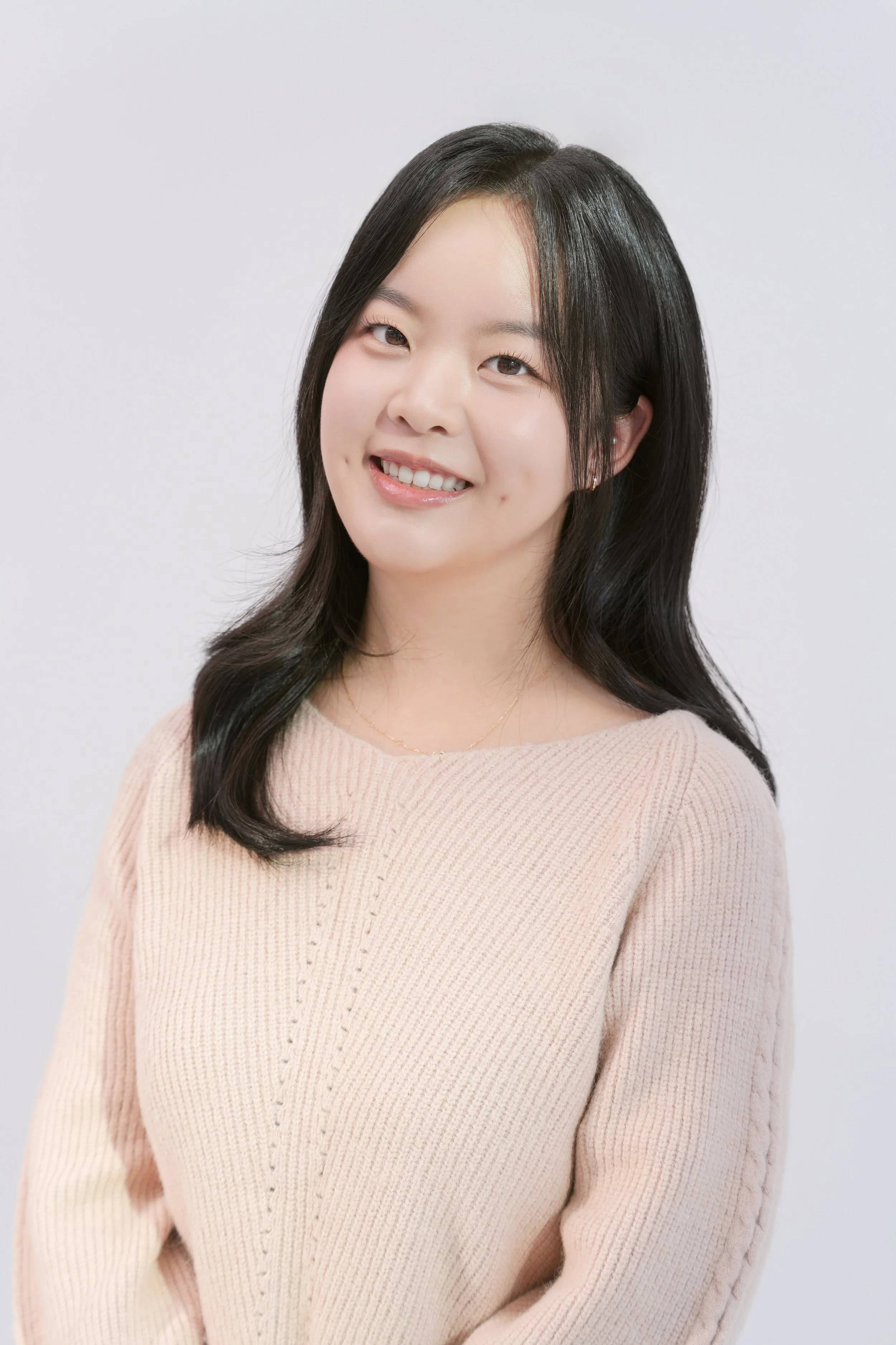 A young woman with long black hair smiling, wearing a beige sweater, against a plain white background.