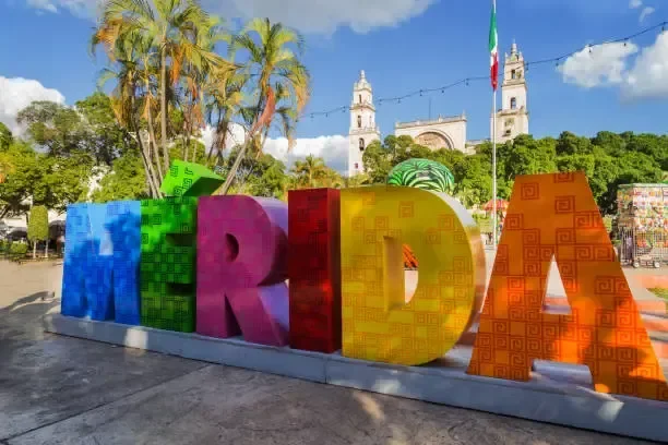 Colorful large 3D letters spelling 'MERIDA' outdoors with trees, a church, and a flag in the background.