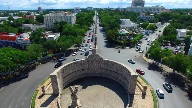 Aerial view of a city street with a monument in the foreground, showing a circular stone structure with sculptures, surrounded by trees and buildings, with traffic moving along the road.
