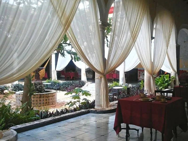 Restaurant dining area with large white curtains, outdoor garden visible through open archways, and a table set with plates and glasses.