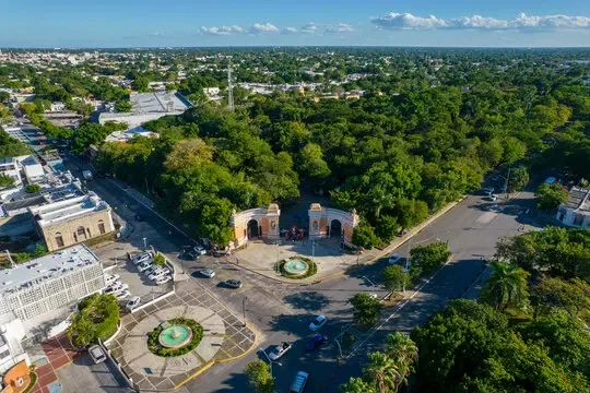 Aerial view of a city park with green trees, a fountain, and surrounding streets with parked cars and buildings under a blue sky.
