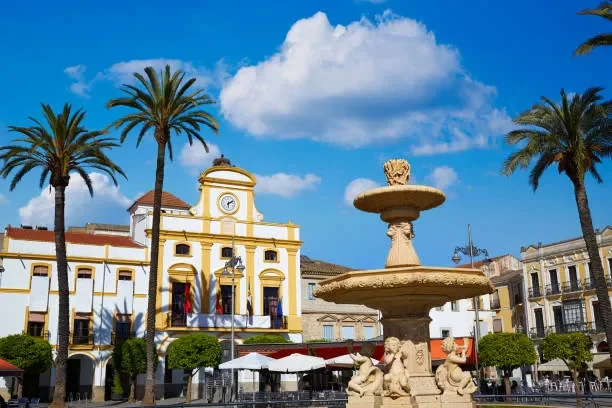 A town square with a yellow and white building, a fountain with lion statues, and palm trees under a partly cloudy blue sky.