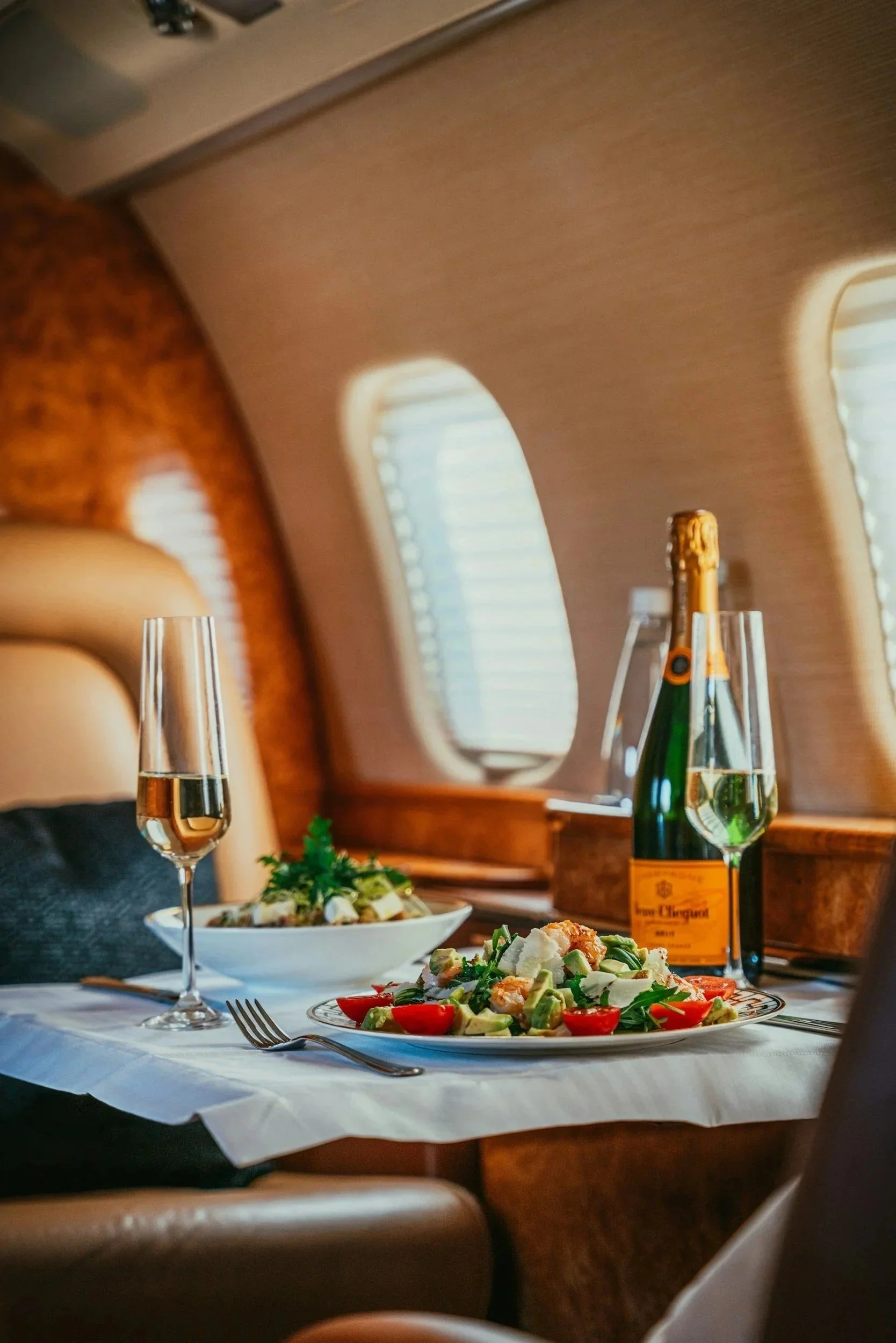 A luxurious meal setup inside an airplane with a glass of champagne, a bottle of champagne, and a plate of salad on a white tablecloth.