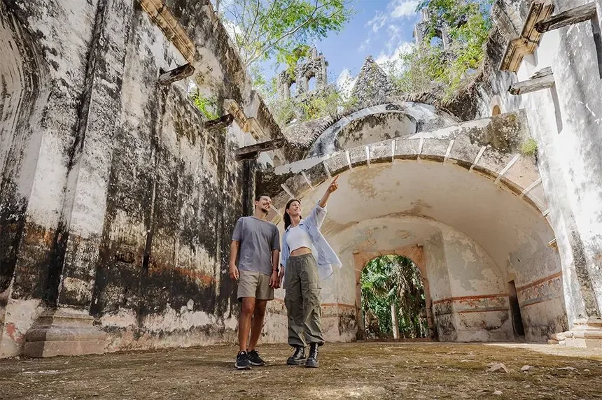 Two people, a man and a woman, standing in front of old, weathered ruins with arches, trees, and blue sky visible in the background. The woman is pointing at something above.