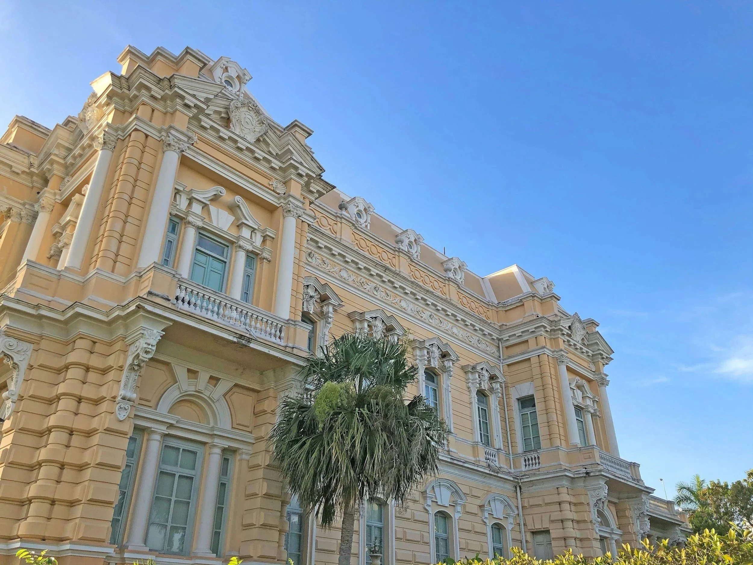 A historic yellow building with white ornate architectural details, tall columns, and arched windows, set against a bright blue sky with a few clouds, partially obscured by a palm tree in the foreground.
