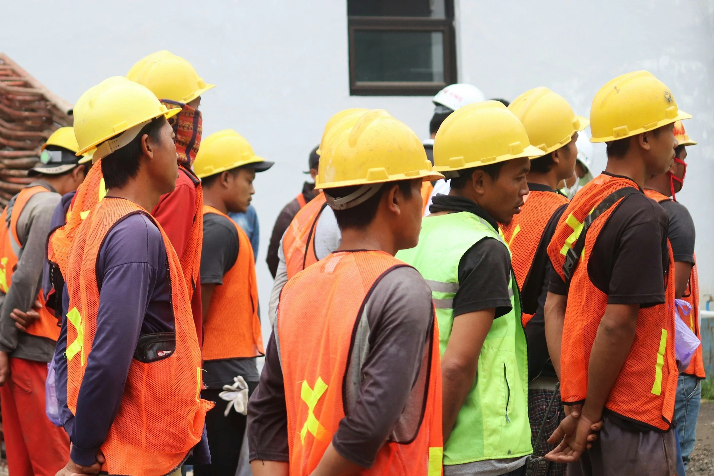 Group of construction workers standing in a line wearing yellow safety helmets and orange safety vests, outdoors near a white wall with a window.