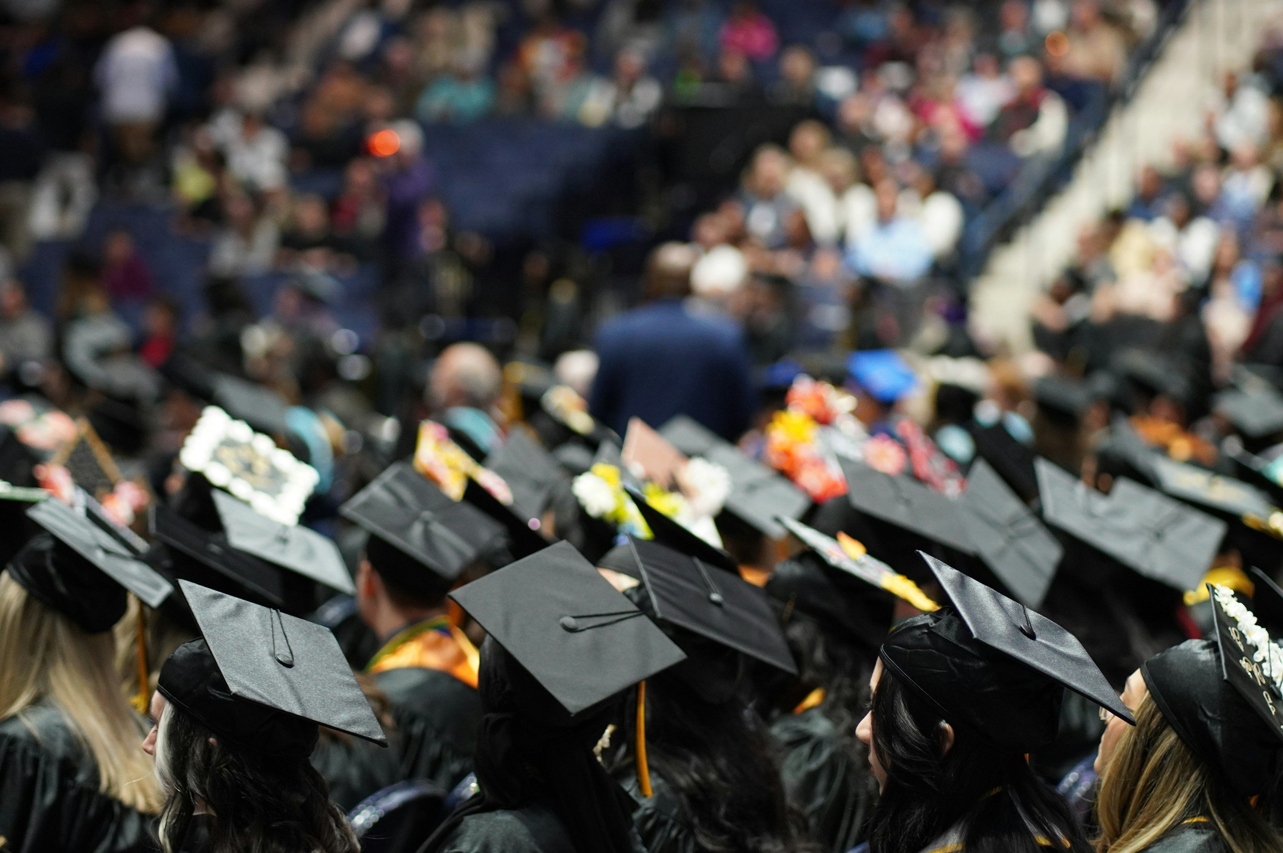 Graduates in caps and gowns at a graduation ceremony, seated in rows with an audience in the background.