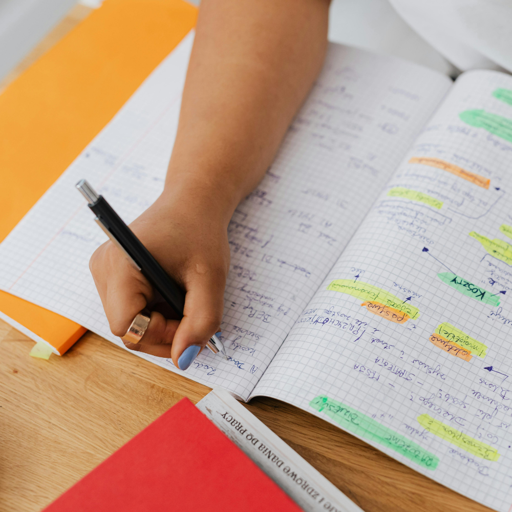 A person's hand with blue painted nails and a silver ring is writing in a graph paper notebook with a black pen. The notebook is open and filled with handwritten notes and highlights. An orange and yellow folder is partially visible in the background, and a red book titled 'Reforming EU Law' is on the desk.