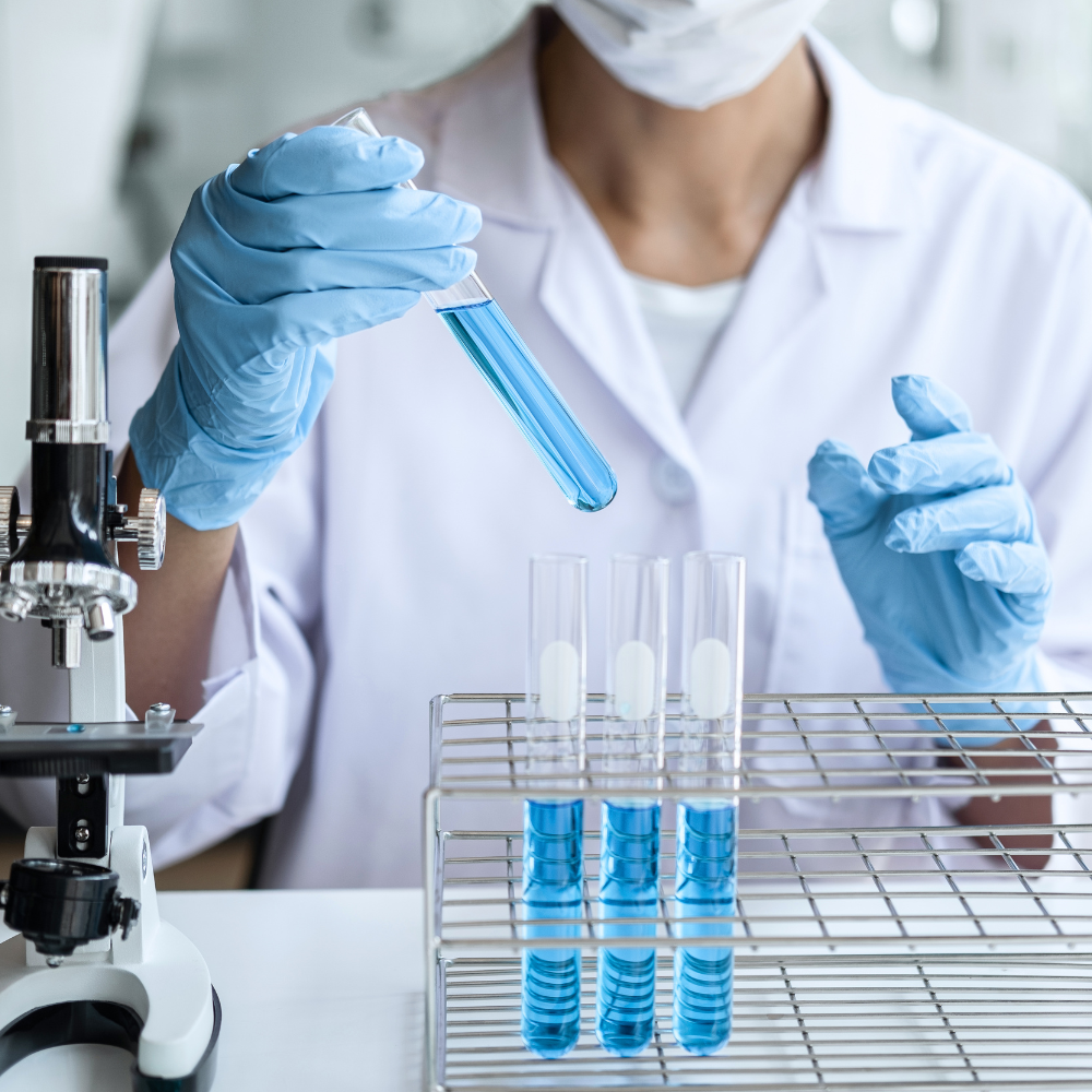 A scientist wearing gloves and a lab coat holds a test tube filled with blue liquid, conducting an experiment in a laboratory with test tubes and a microscope.