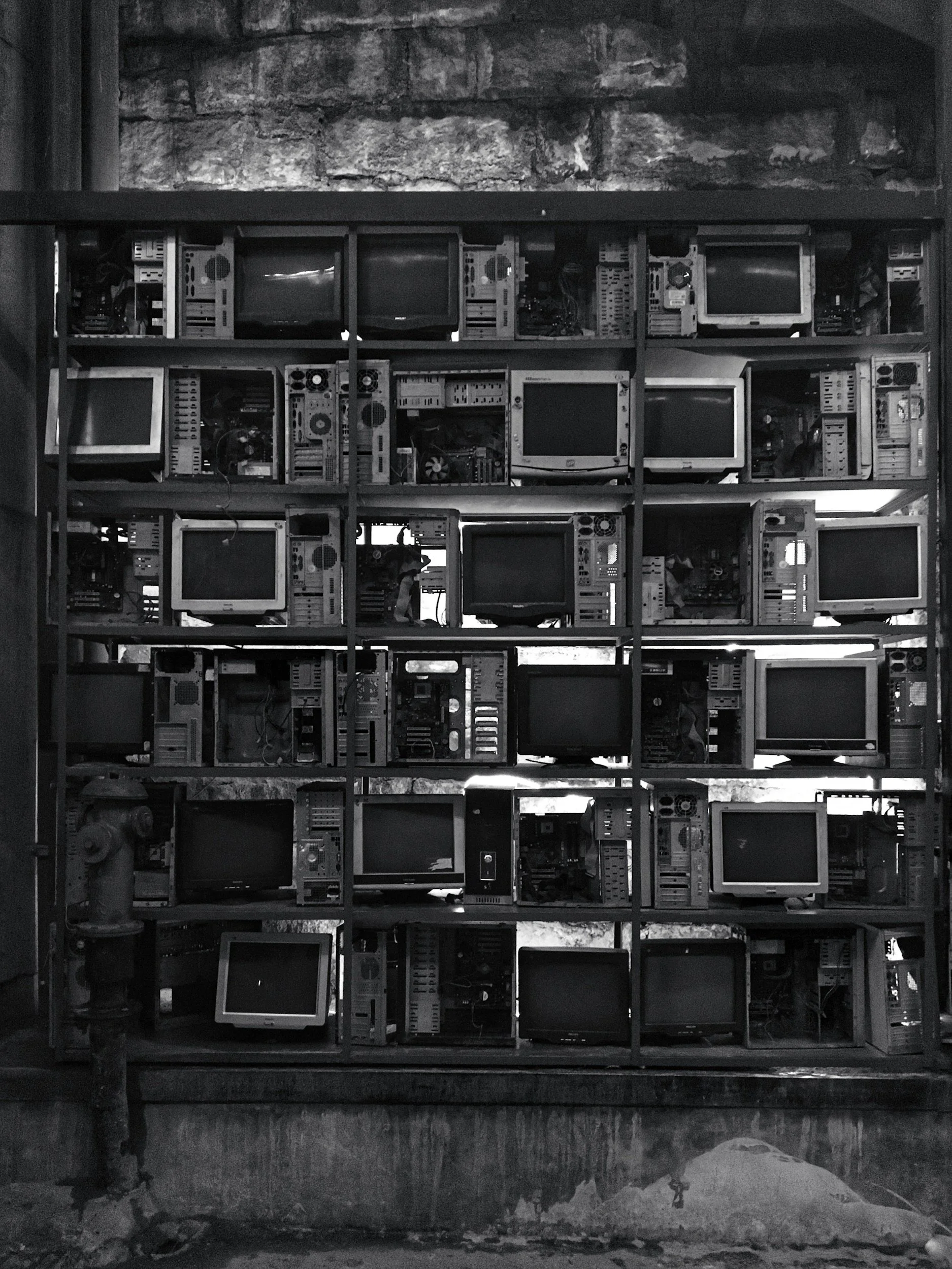 A metal grid shelf holding multiple old CRT monitors and computer parts, arranged in a grid pattern with some gaps, against a brick wall background.