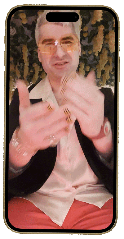 Person wearing glasses and stylish attire, gesturing with hands, in a dimly lit environment with leafy décor.