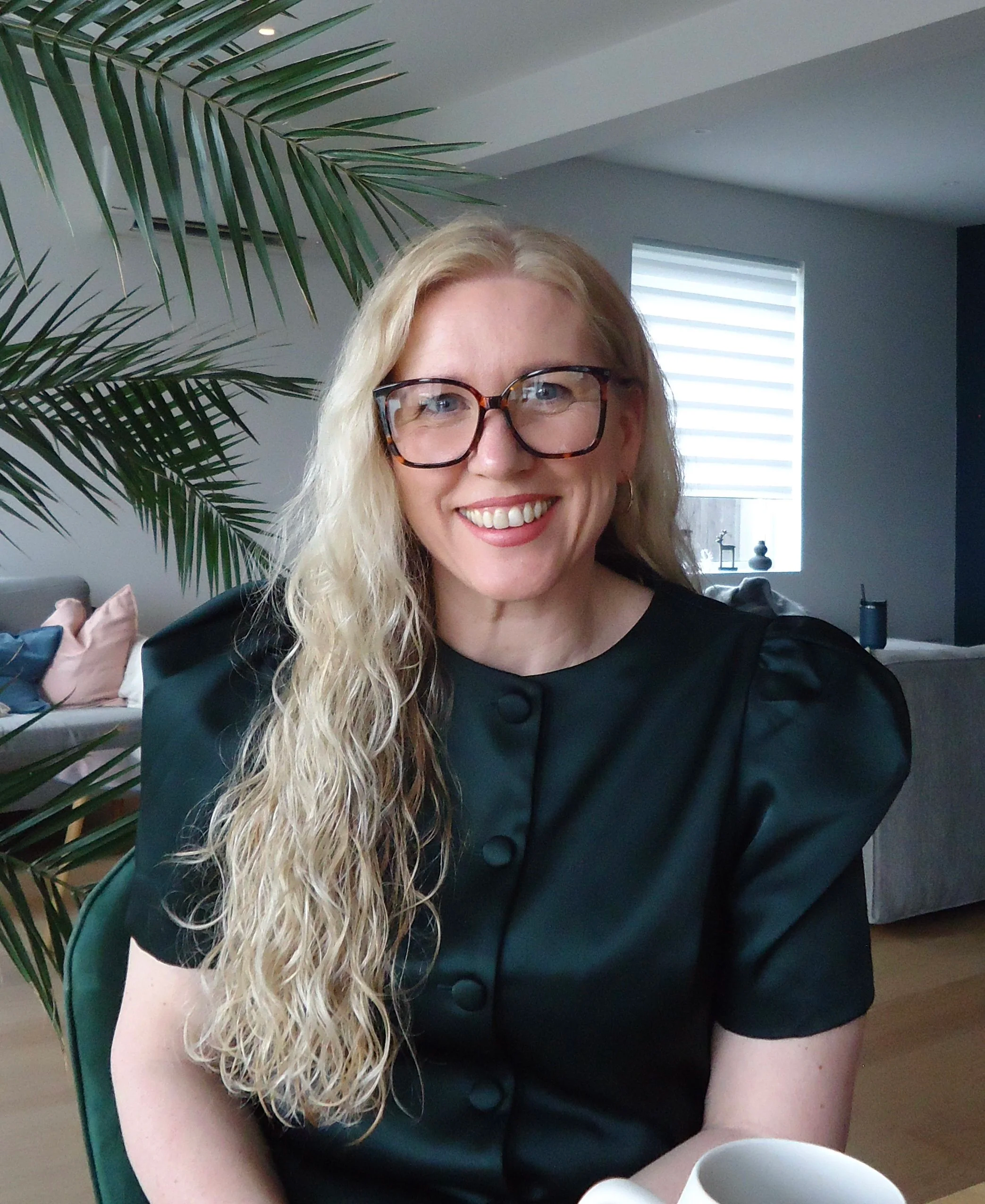 En kvinne med langt, krøllete blondt hår, glade smil, store rektangulære briller, svart kjole med knappestil, sitter ved et bord med en hvit kopp i en moderne stue med store vinduer, grønne planter og sofaputer.