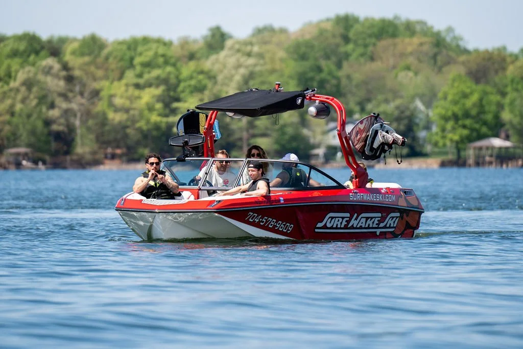 A red and black wakeboarding boat on a calm lake with four people on board, surrounded by green trees under a blue sky.