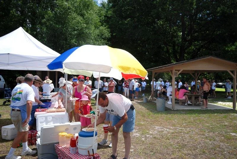 Crowd gathered at a park pavilion during a community event with people serving drinks