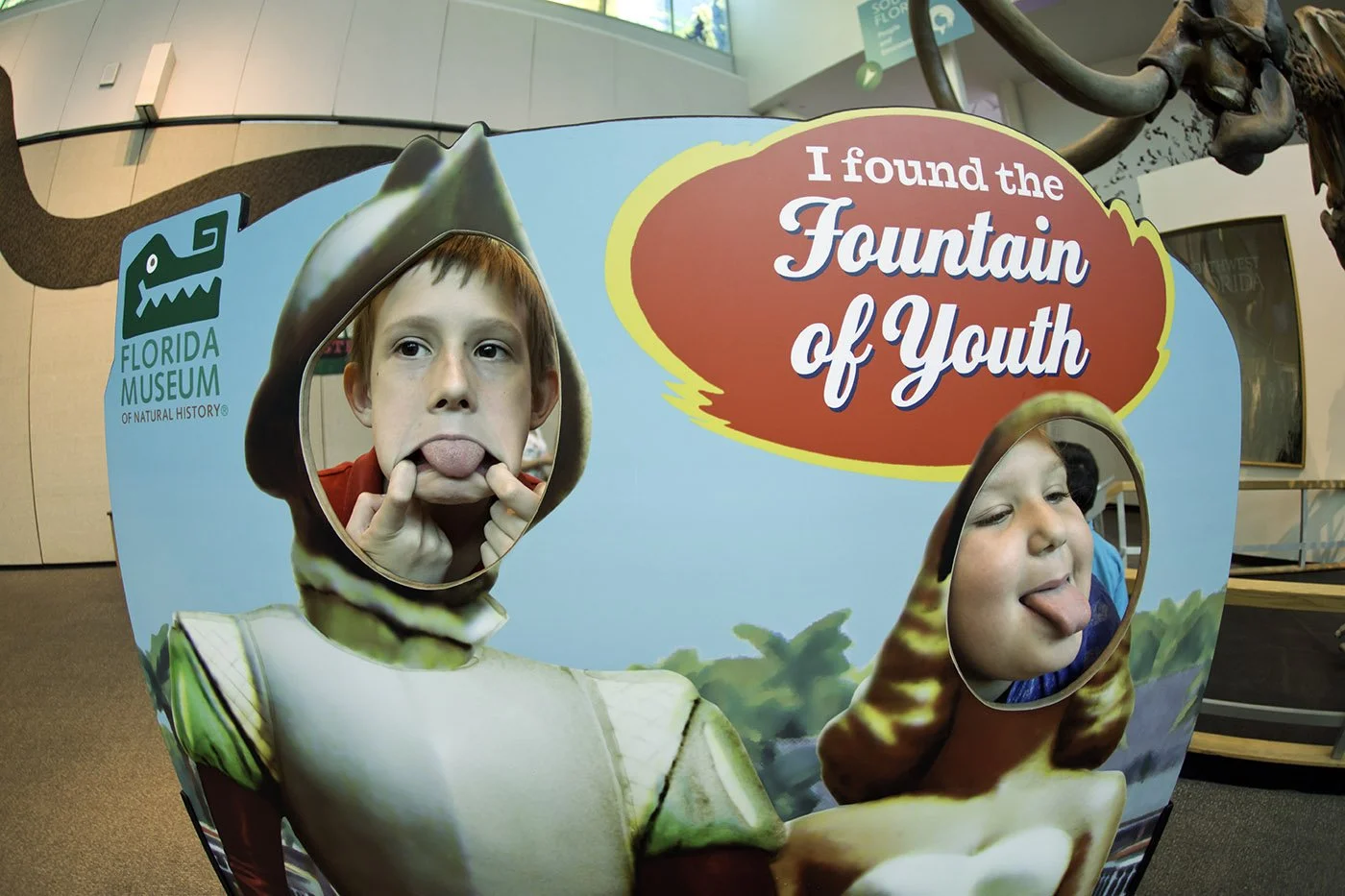 Two children pose with their faces in a cutout display about the Fountain of Youth at the Florida Museum of Natural History, making playful expressions
