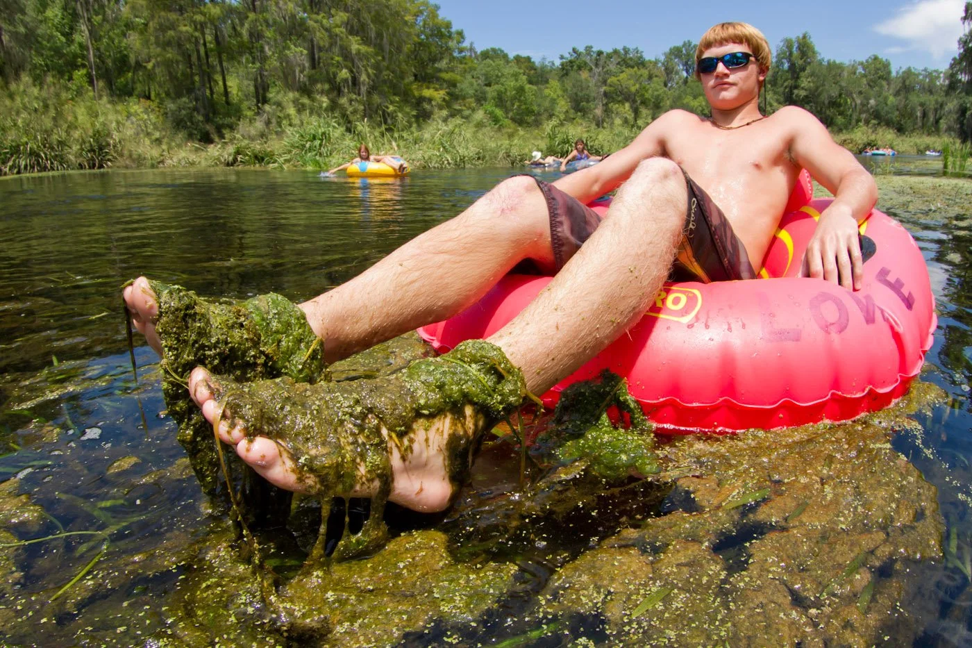 Person relaxing in an inner tube on a clear spring, with feet covered in algae and aquatic vegetation visible beneath the water