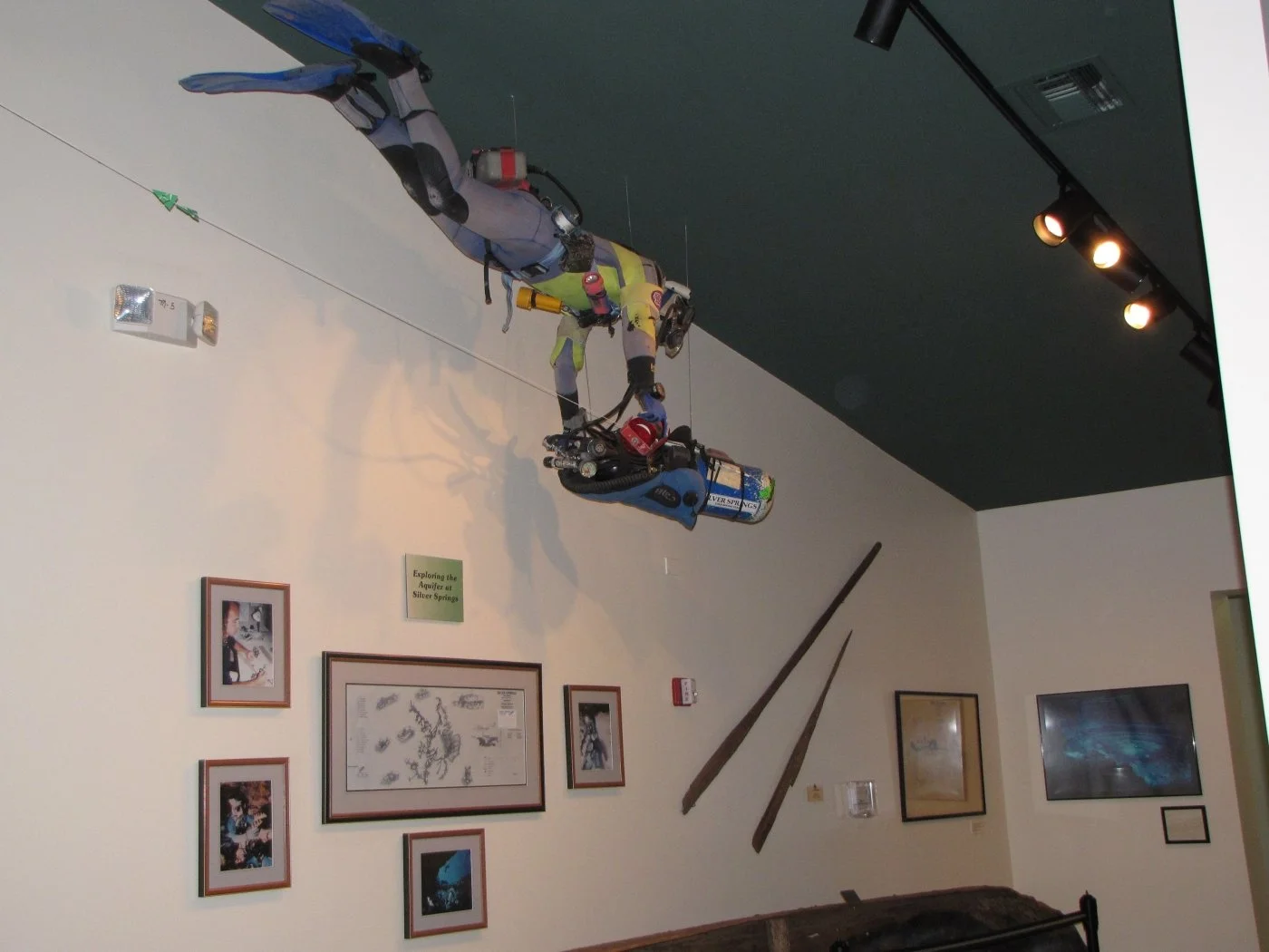 Diving equipment suspended from the ceiling in a museum exhibit, with framed photos and displays on the surrounding walls.