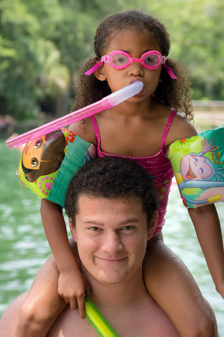 Child wearing pink swim goggles and floaties sits on an adult’s shoulders in the water, holding a snorkel and looking forward