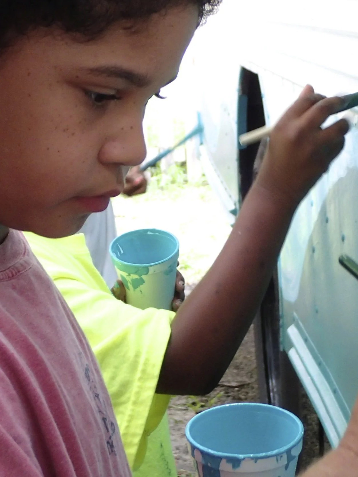 Close-up of a child carefully painting a bus surface while holding a cup of paint.