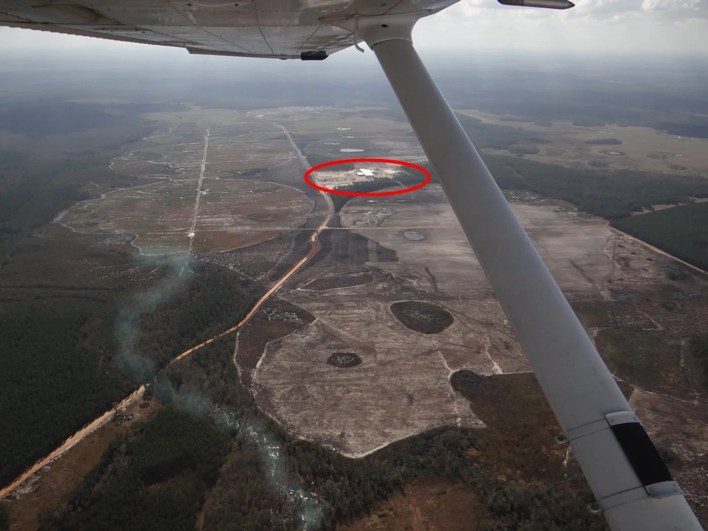 Aerial view of cleared land and forest in Putnam County, Florida, taken from a small plane. A white building in the distance is circled in red, and a creek is visible in the foreground running through the trees.