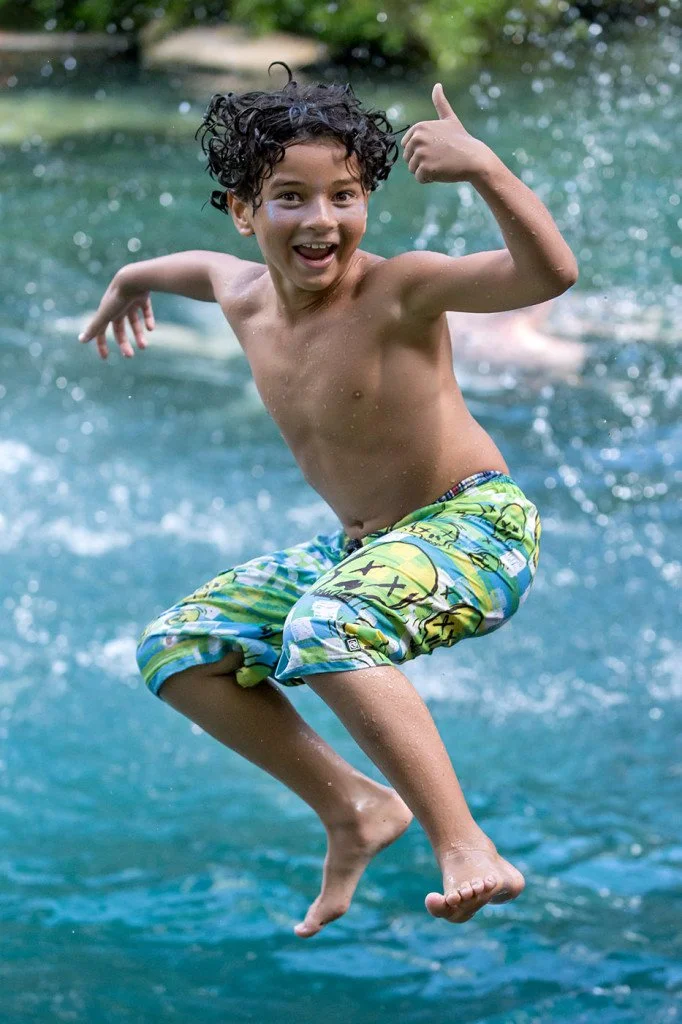 Child jumping into clear blue spring water with a joyful expression, giving a thumbs up while splashing midair.