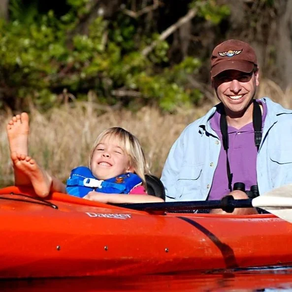 Child reclining and smiling in a red kayak while an adult sits behind them on calm water