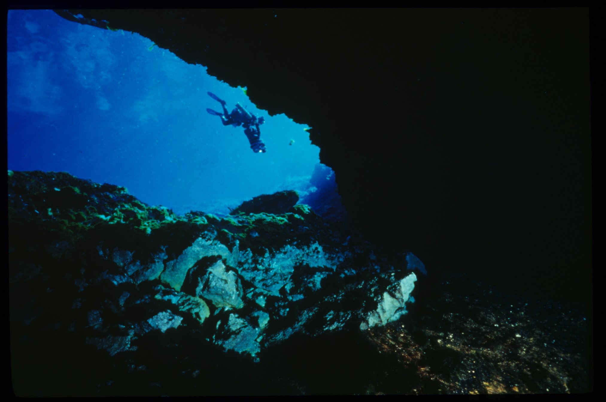 Scuba diver entering a dark underwater cavern with blue spring light visible beyond rocky formations