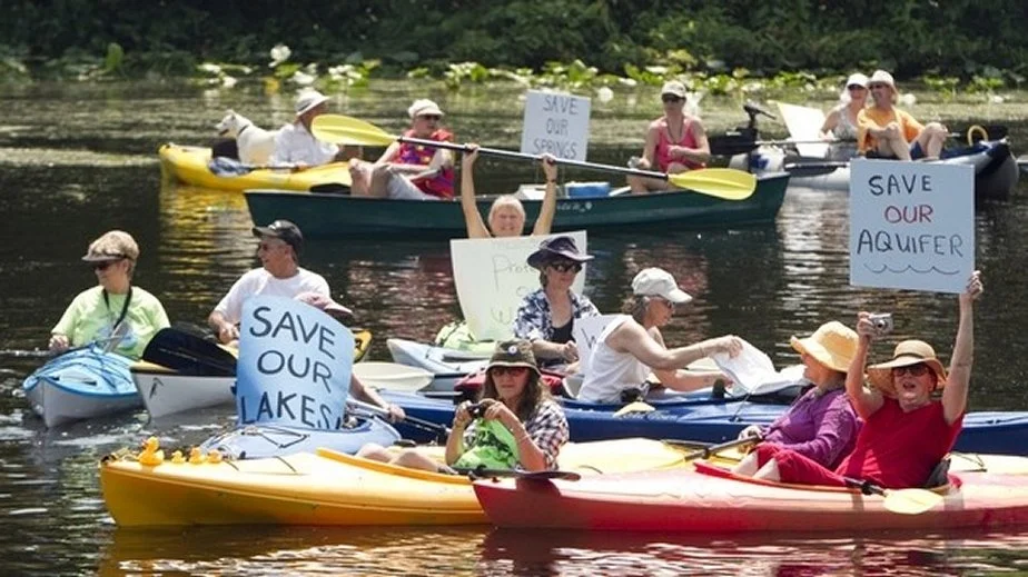 Group of people in kayaks and canoes holding signs reading “Save Our Lakes,” “Save Our Springs