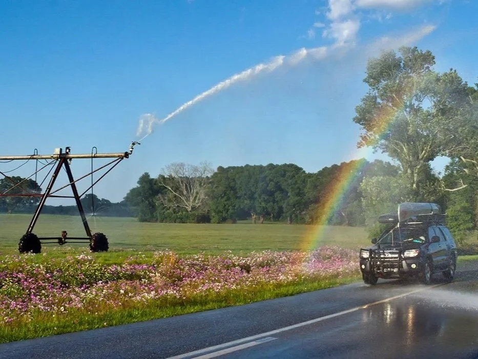 Center pivot irrigation spraying water across a field, creating a rainbow over a roadside landscape.