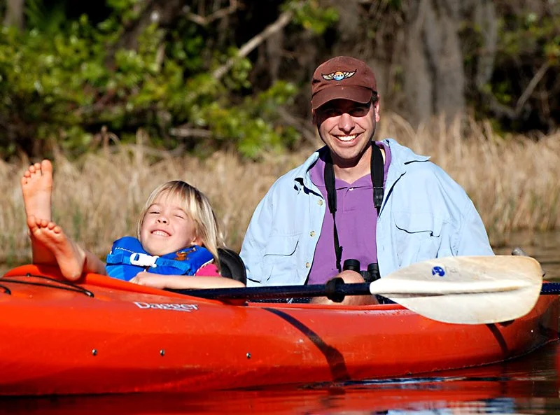 Child reclining and smiling in a red kayak while an adult sits behind them on calm water