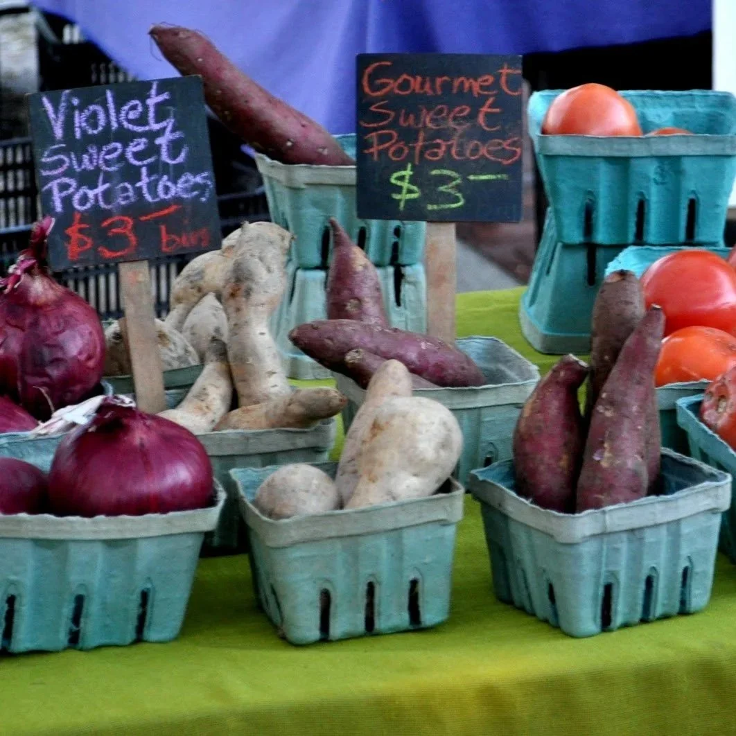 Baskets of sweet potatoes, onions, and tomatoes displayed at a farmers market with handwritten price signs