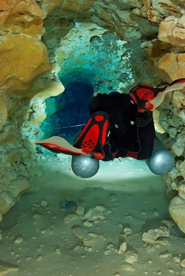 Scuba diver exploring an underwater cave tunnel with rocky walls and sandy floor in a spring system