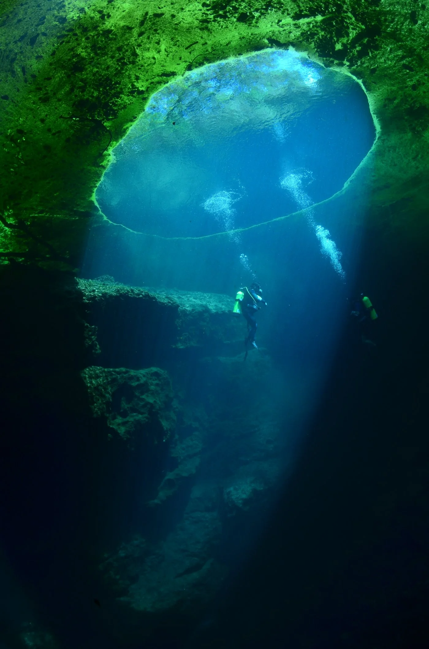 Scuba divers descending into a deep underwater spring opening with sunlight streaming through the surface above.