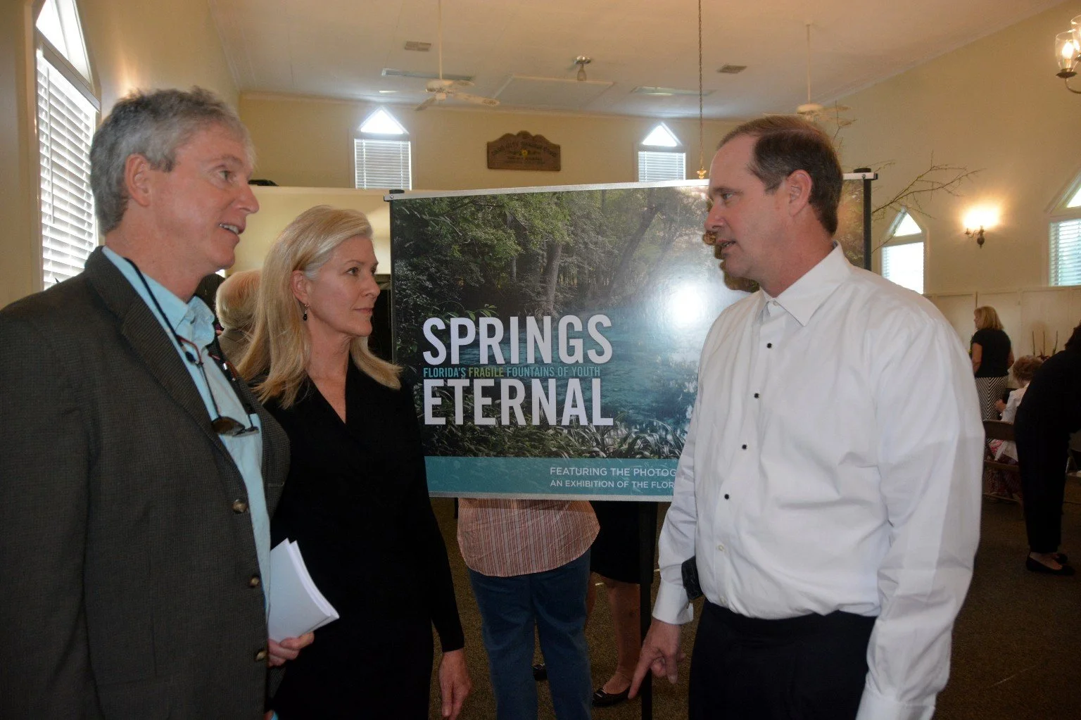 People speaking together at a Springs Eternal exhibition event, standing beside a display sign about Florida springs conservation.