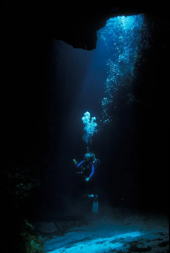 Scuba diver descending in a dark underwater cavern with a beam of blue light and rising air bubbles.