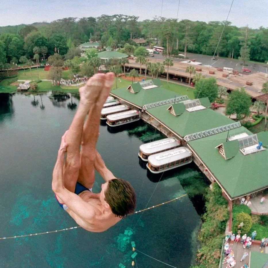 Diver jumping from a high platform into clear water at Silver Springs with docks and boats nearby.