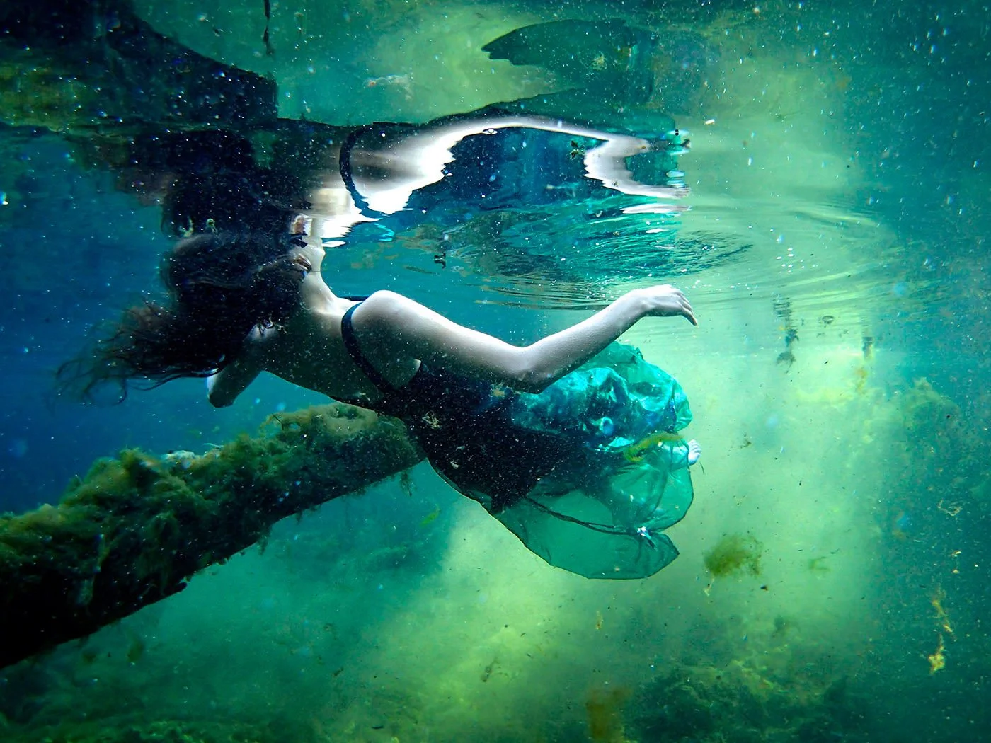 A person floats just beneath the surface of a freshwater spring, their body partially reflected above the waterline. A sheer green fabric trails behind them, with a submerged log and suspended particles visible in the water.