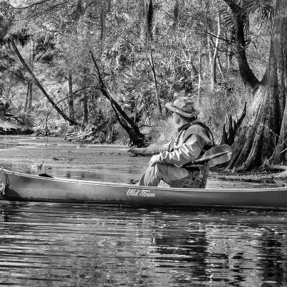 Black-and-white photograph of Lars Andersen sitting in a canoe on the Ichetucknee River, surrounded by trees along the riverbank.