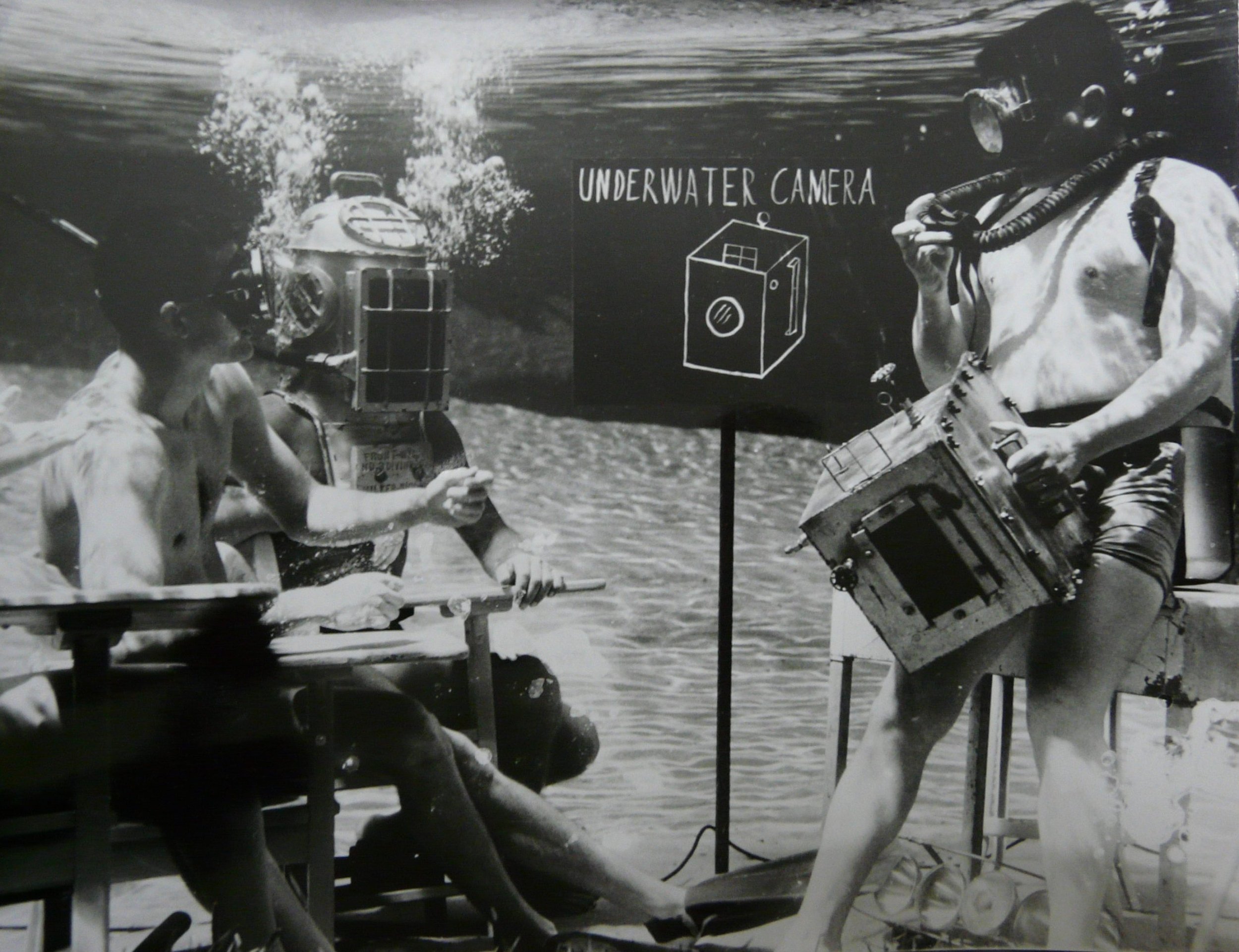 Underwater photograph by Bruce Mozert showing a diver using a large camera while another person sits nearby, with a sign reading “Underwater Camera” in the background.