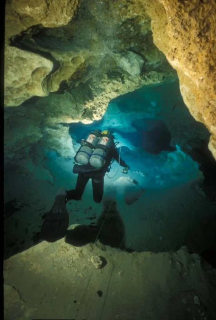 Scuba diver swimming through a submerged limestone cave with clear blue water and rock formations