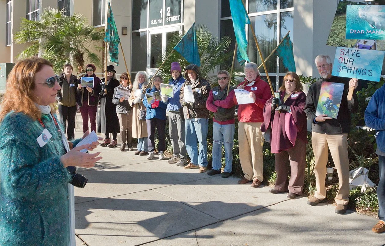 A speaker addresses a group of environmental advocates gathered outside a building during a clean water rally, while participants hold signs and images supporting the protection of Florida’s springs.