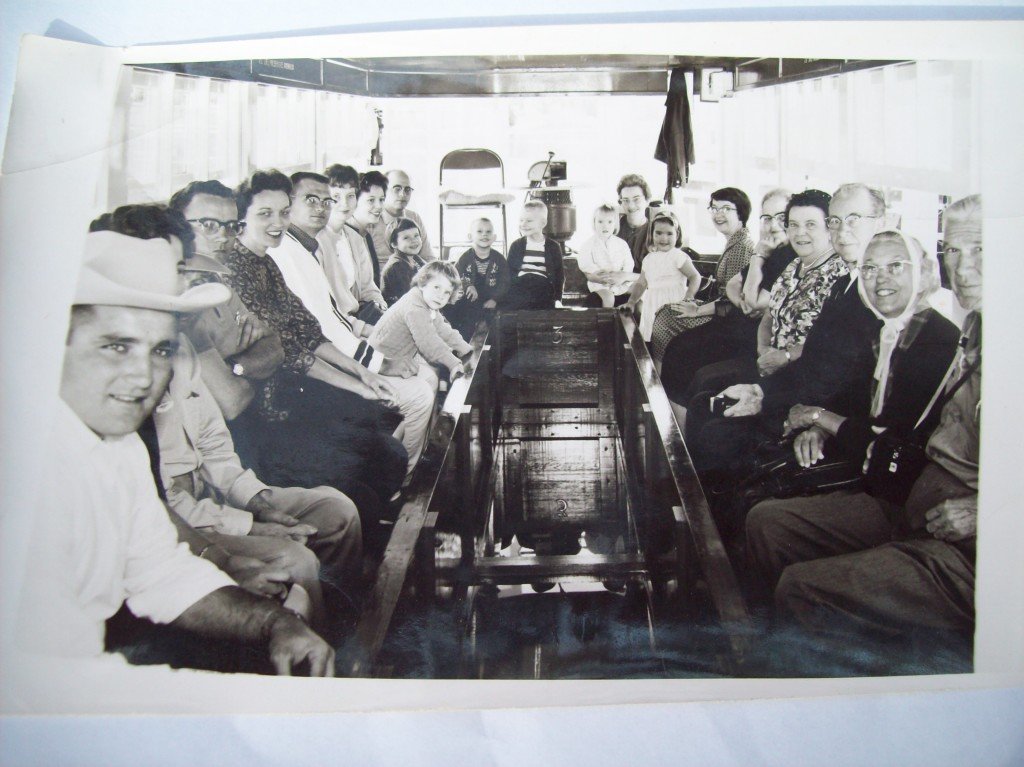 Historic black-and-white photo of a group of adults and children seated on benches inside a glass-bottom boat, looking down into the water below.