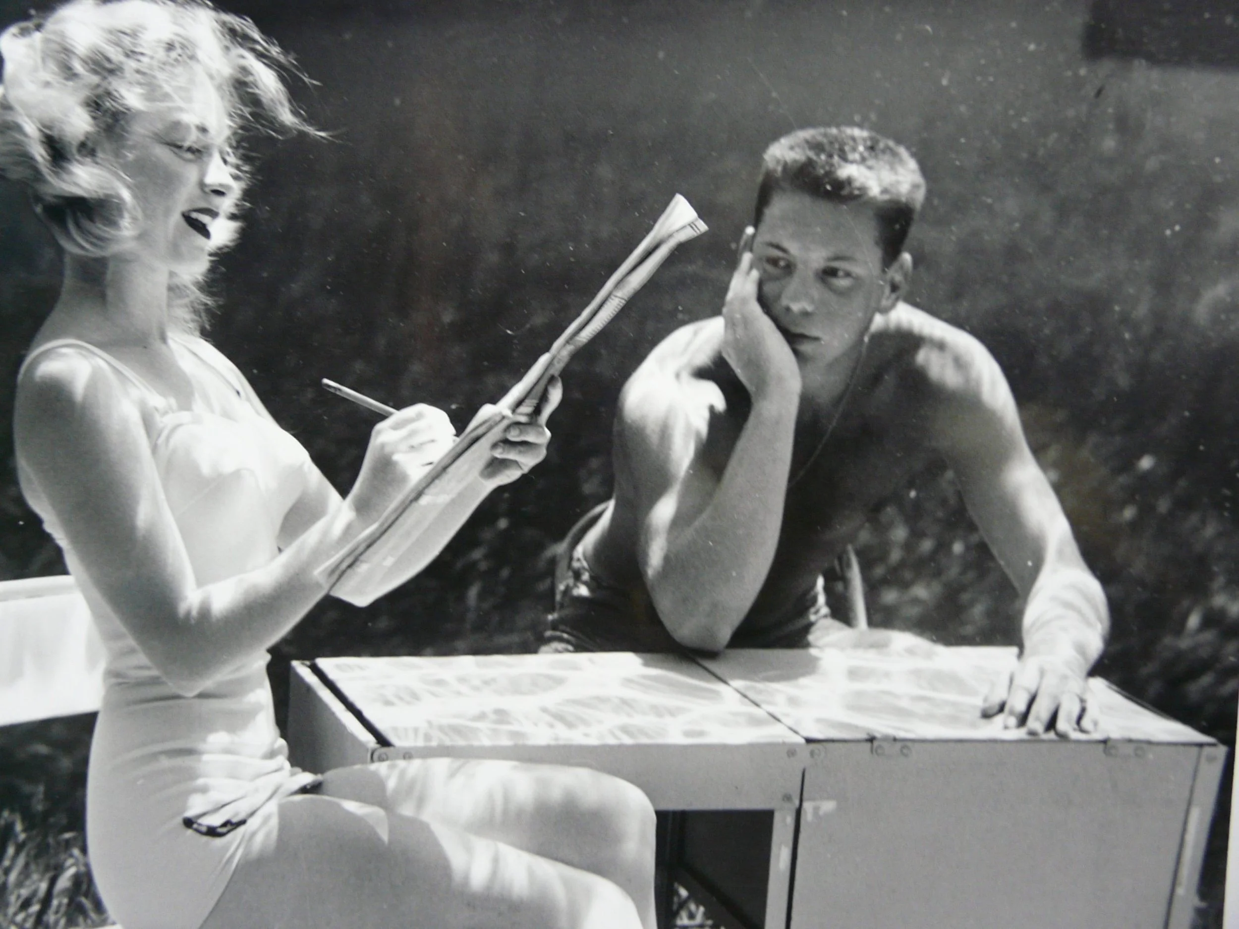 Underwater photograph by Bruce Mozert of a woman in a dress writing on a pad at a table while a man sits beside her, creating a staged, humorous scene.