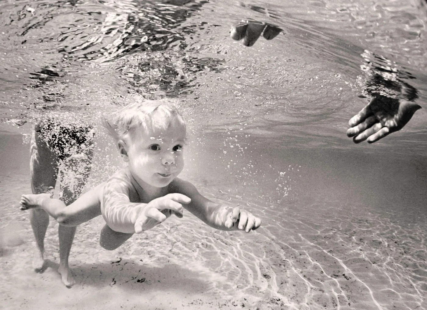 Black and white underwater photo of a baby swimming at Poe Springs with an adult's legs visible behind them"Black and white underwater photo of a baby swimming at Poe Springs with an adult's legs visible behind them.