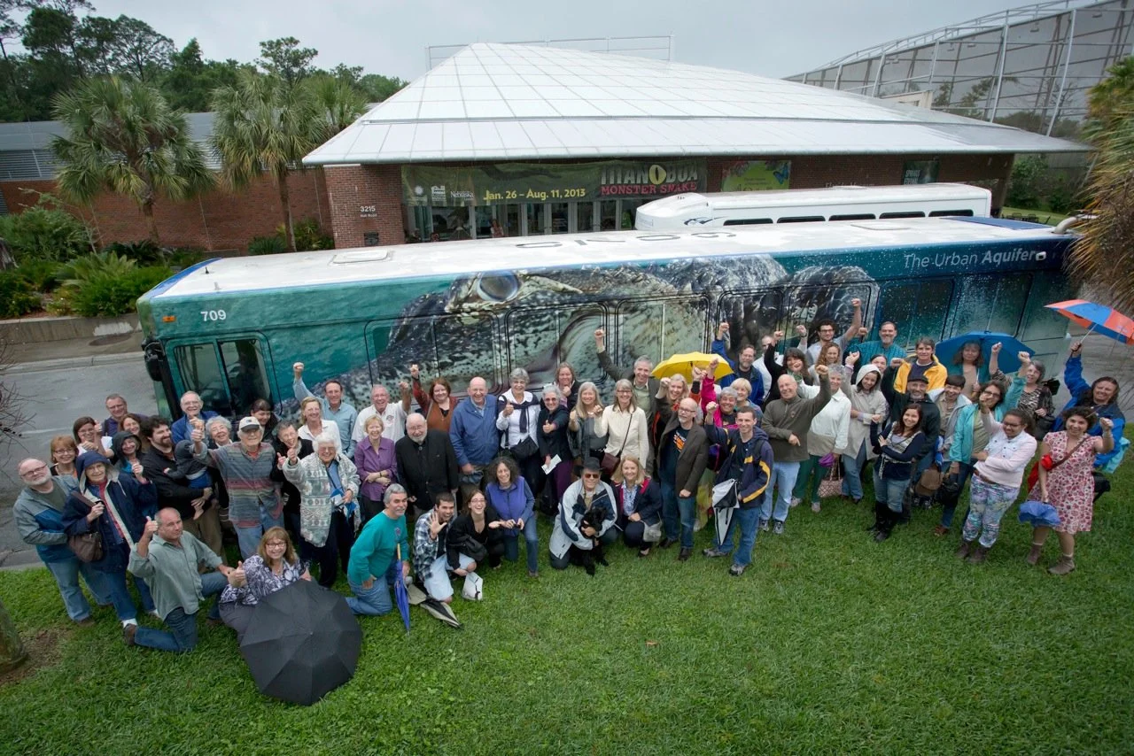 Group of people gathered in front of a city bus wrapped with spring imagery for the Urban Aquifer public art project outside a museum building.
