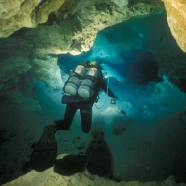 Scuba diver with twin air tanks swimming through a limestone underwater cave, illuminated by blue spring water filtering through the cavern.