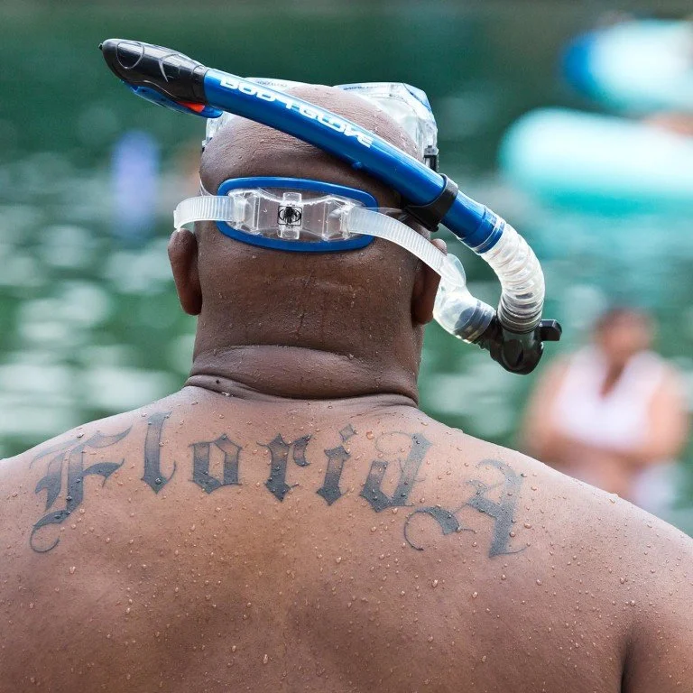 Image of the back of a man's head wearing a snorkel mask and upper back with the word "Florida" tattooed across his shoulders in a gothic style.